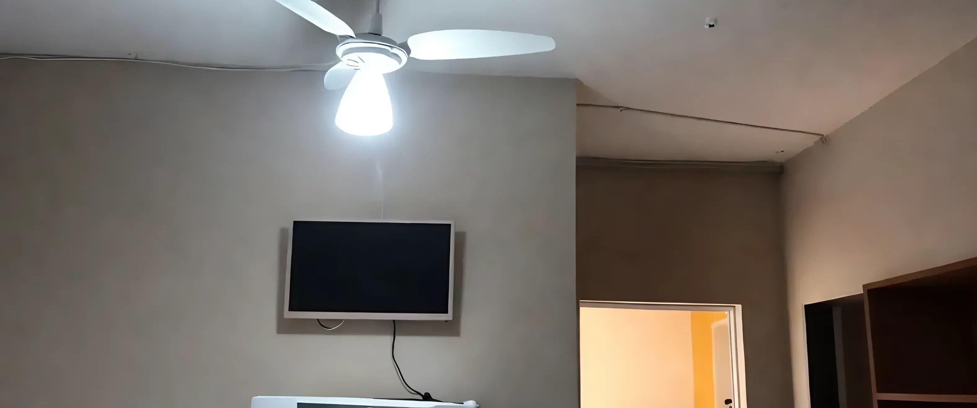 A ceiling fan with a light fixture, a mounted flat-screen TV, and a doorway leading to another room.