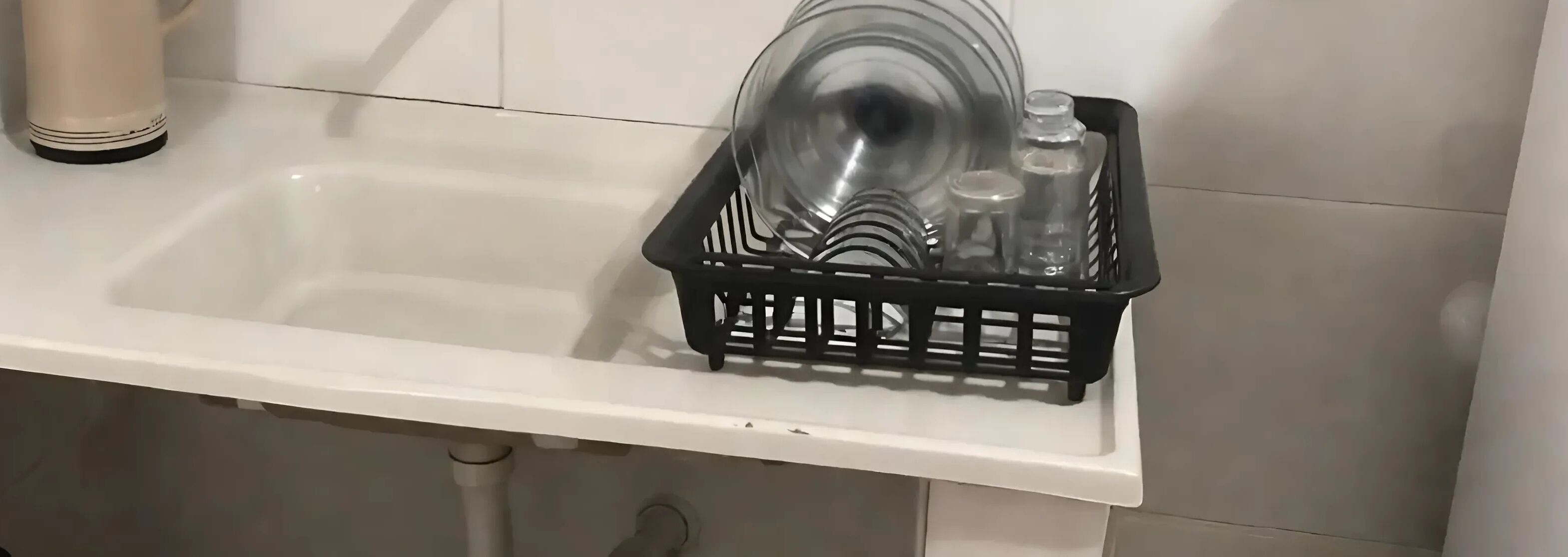 Un lavamanos de cocina con un escurreplatos negro que contiene cristalería transparente y una tetera blanca.