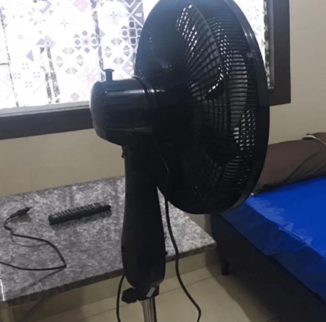 A room with a standing fan, a remote control, and a bed with a blue cover.