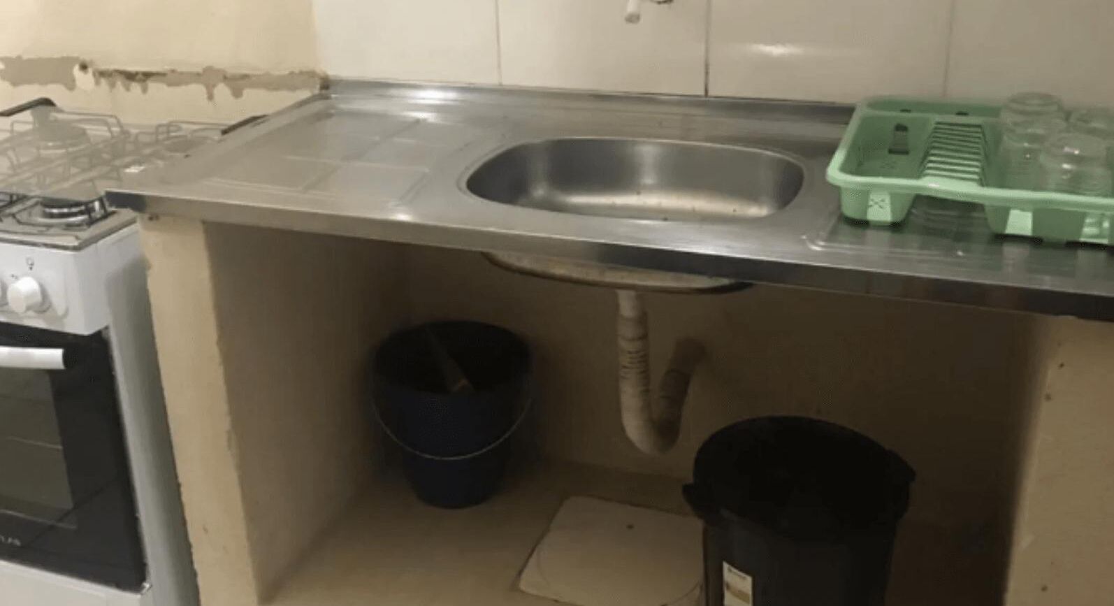 Una cocina con un lavamanos de acero inoxidable, una estufa a gas, un cubo azul y un bote de basura negro.