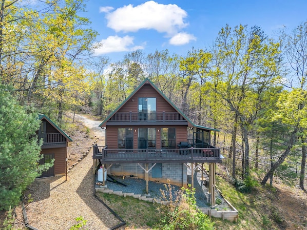 A cabin with a balcony, a covered deck, and a gravel driveway.