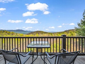 Una terraza con una mesa de vidrio y sillas de metal, con vistas a un paisaje boscoso y montañas a lo lejos.