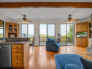 A spacious living area with a wooden floor, a blue sofa, a dining table, and a kitchen with wooden cabinets and a large window.