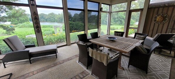 Un solárium con un amplio ventanal que da a un jardín y un lago. Tiene una mesa de madera con sillas y un pequeño sofá.
