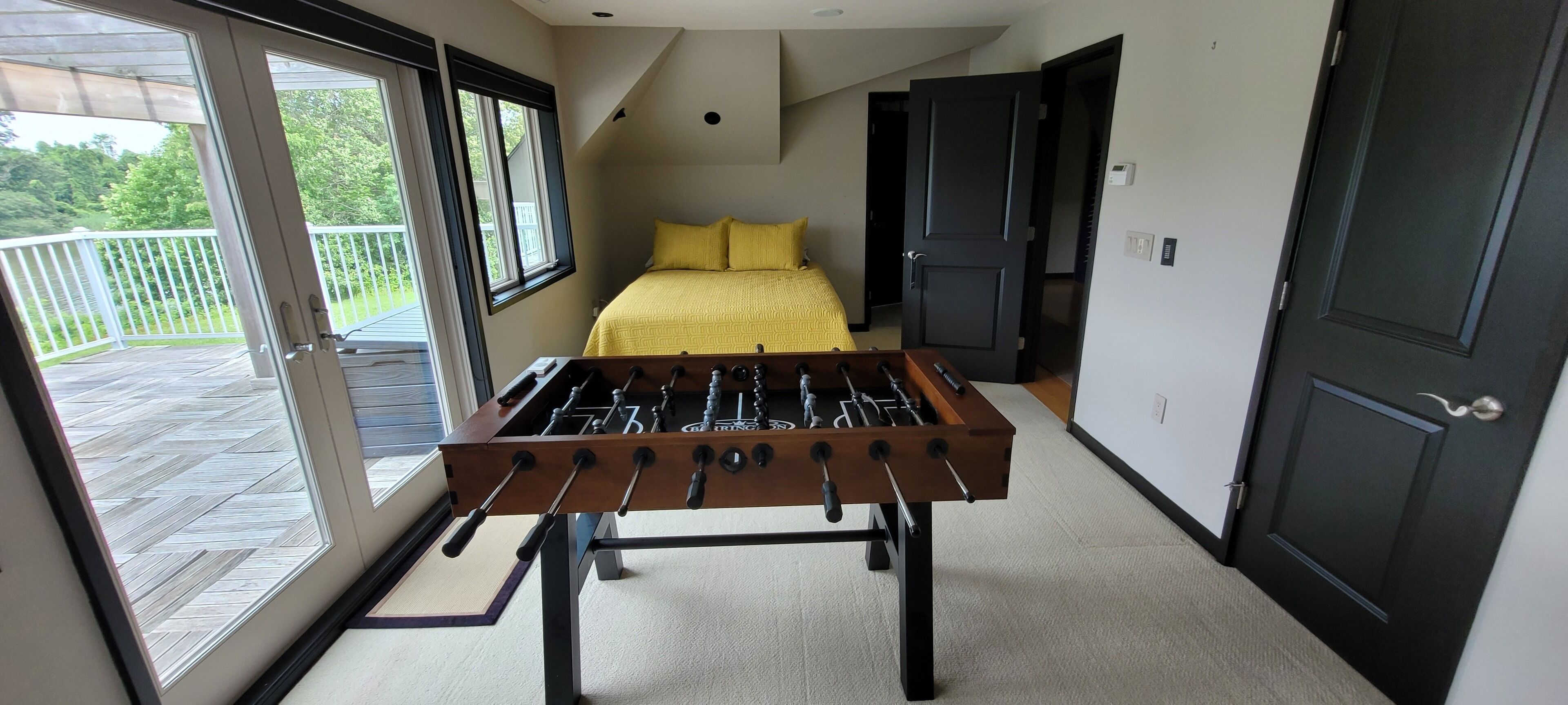 A room with a foosball table, a bed with yellow bedding, and a balcony with a wooden deck.