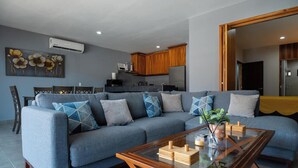 Un salon moderne avec un canapé gris, une table basse en bois et un coin repas, la cuisine étant visible au fond.