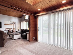 Ein großzügiges Wohnzimmer mit hoher Decke, Kamin, Essbereich mit Stühlen und großen Fenstern mit Jalousien.