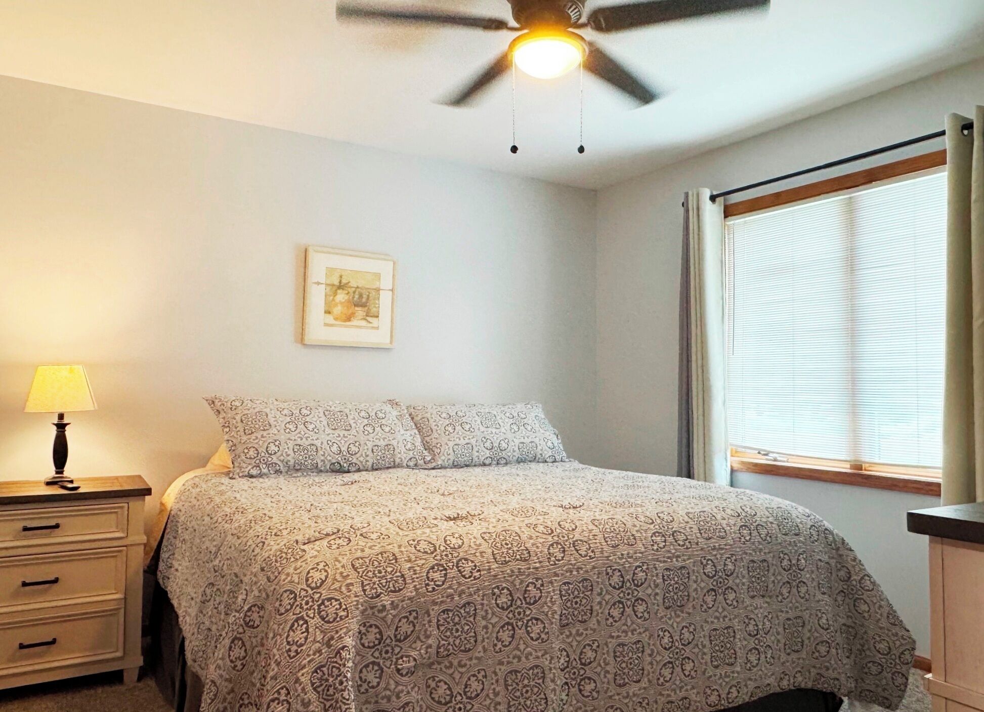 A bedroom with a bed, a nightstand, a lamp, a ceiling fan, a window with blinds, and a framed picture on the wall.