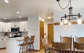A modern kitchen with white cabinets, a microwave, and an island with chairs. There is a dining area with a wooden table and chairs. - Gun lake 4 Bed 2.5 Bath Home w/2 car garage, kayaks, paddleboards, free Wi-Fi (Wayland)