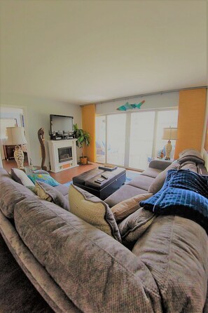 A living room with a large sofa, a coffee table, and a television. There is a plant and a window with curtains.