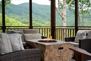 Uma área de estar coberta ao ar livre com lareira, móveis de vime e vista para uma montanha arborizada.