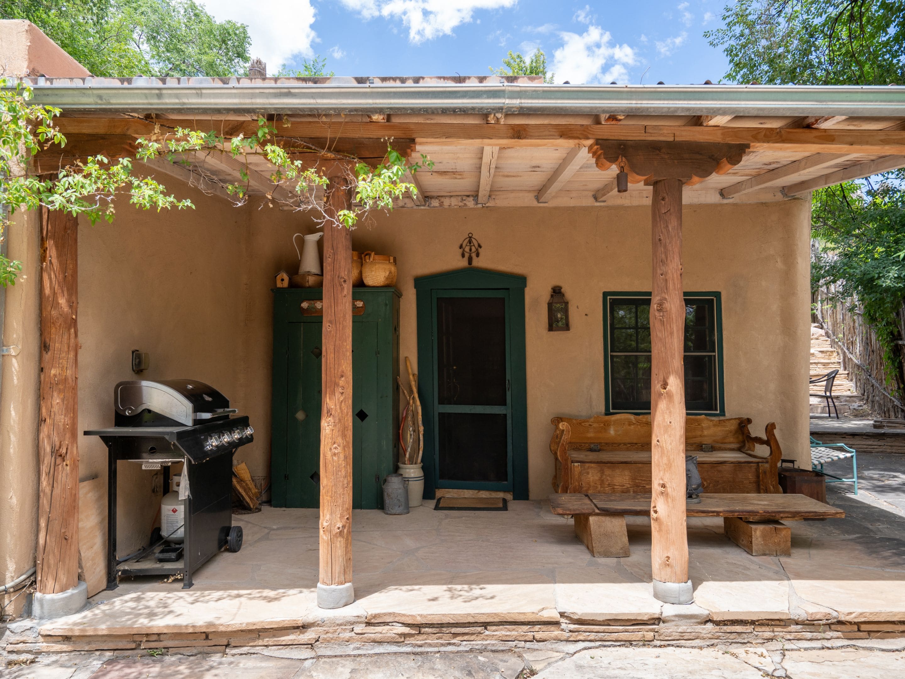 A rustic outdoor patio with a wooden grill, a green door, and a wooden bench.