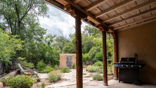 Un patio couvert avec un sol en pierre, des piliers en bois, un barbecue et, en arrière-plan, un bâtiment au style rustique.