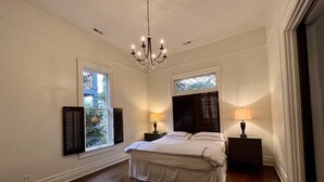 A bedroom with a large bed, two bedside tables, a chandelier, and two windows with shutters.
