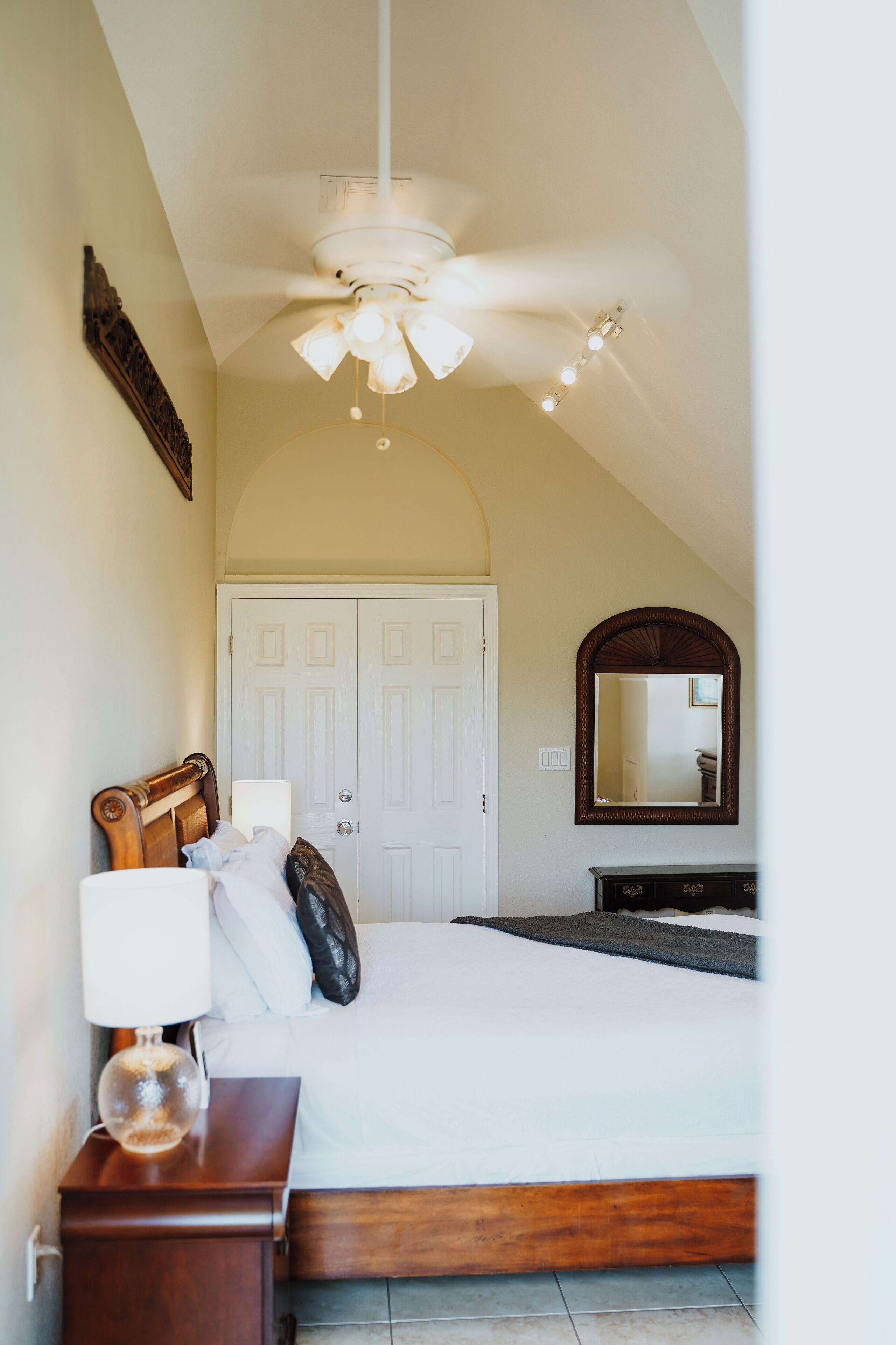 Une chambre à coucher comprenant un lit, une table de chevet, une lampe, un ventilateur de plafond, une armoire et un miroir.