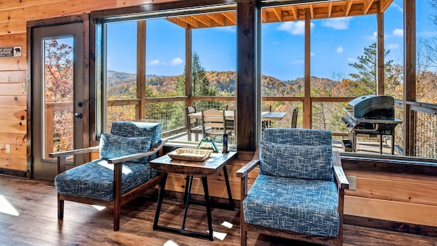 Une cabane en bois dotée de grandes fenêtres qui offrent une vue sur un paysage montagneux. À l’intérieur, on trouve deux fauteuils bleus recouverts de tissu et une petite table sur laquelle est posé un plateau.