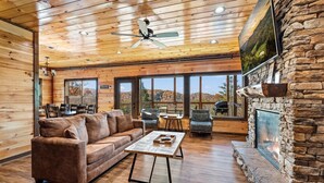 L’intérieur chaleureux d’un chalet avec un plafond en bois, une cheminée en pierre, une grande fenêtre donnant sur un paysage magnifique et un coin salon confortable.