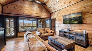 L’intérieur chaleureux d’un chalet avec un plafond en bois, de grandes fenêtres, un canapé, une table basse et un téléviseur fixé au mur.