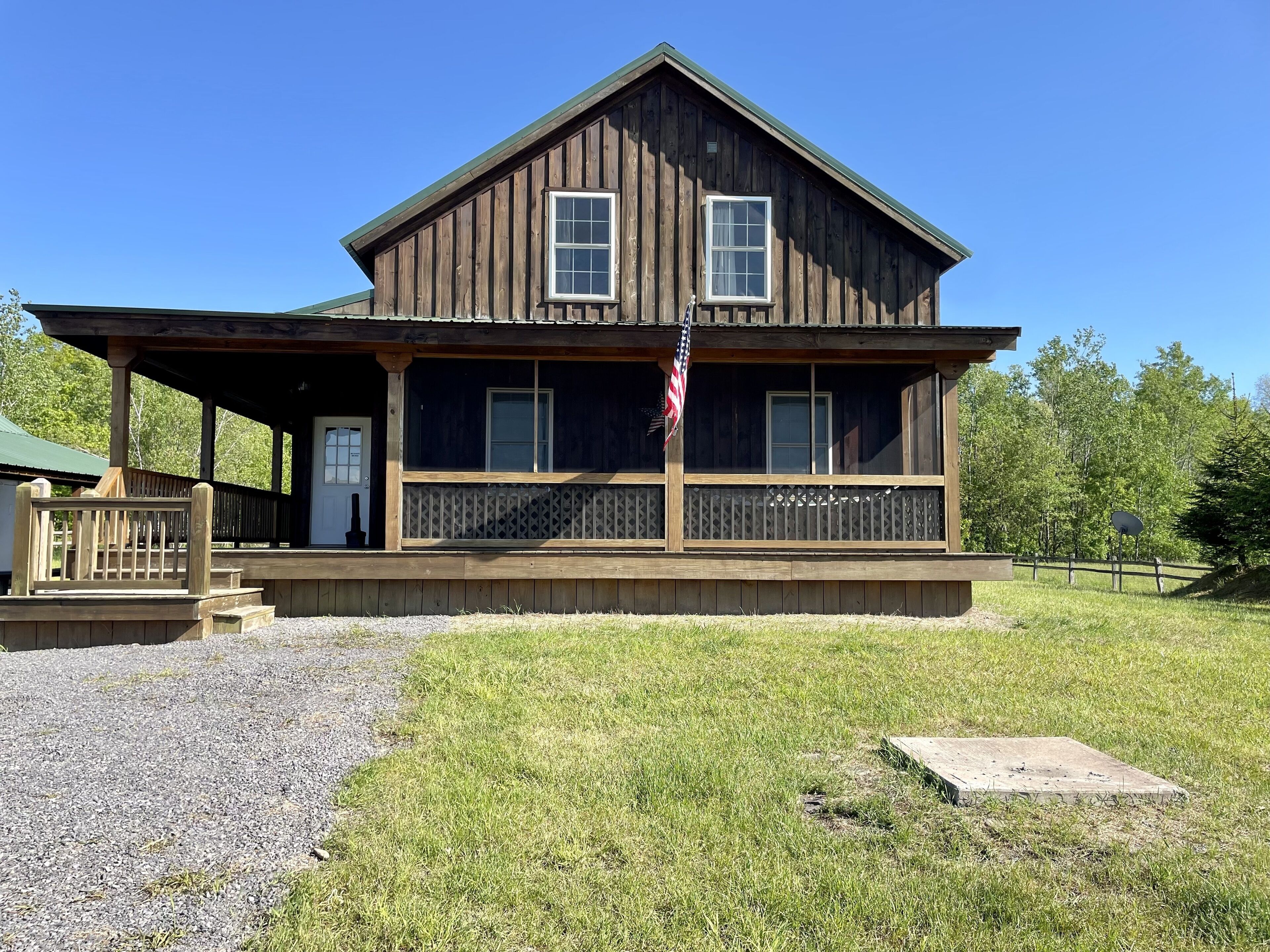 A two-story wooden cabin with a porch, a flag, and a gravel driveway.
