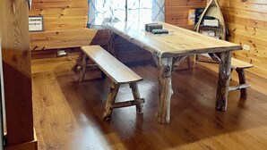 A wooden cabin interior with a large dining table, benches, and a hanging light fixture.