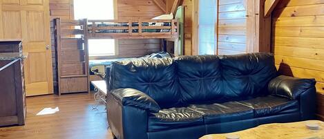 L’intérieur chaleureux d’un chalet avec un plafond en bois, un canapé en cuir noir, une table basse en bois et un lit superposé.