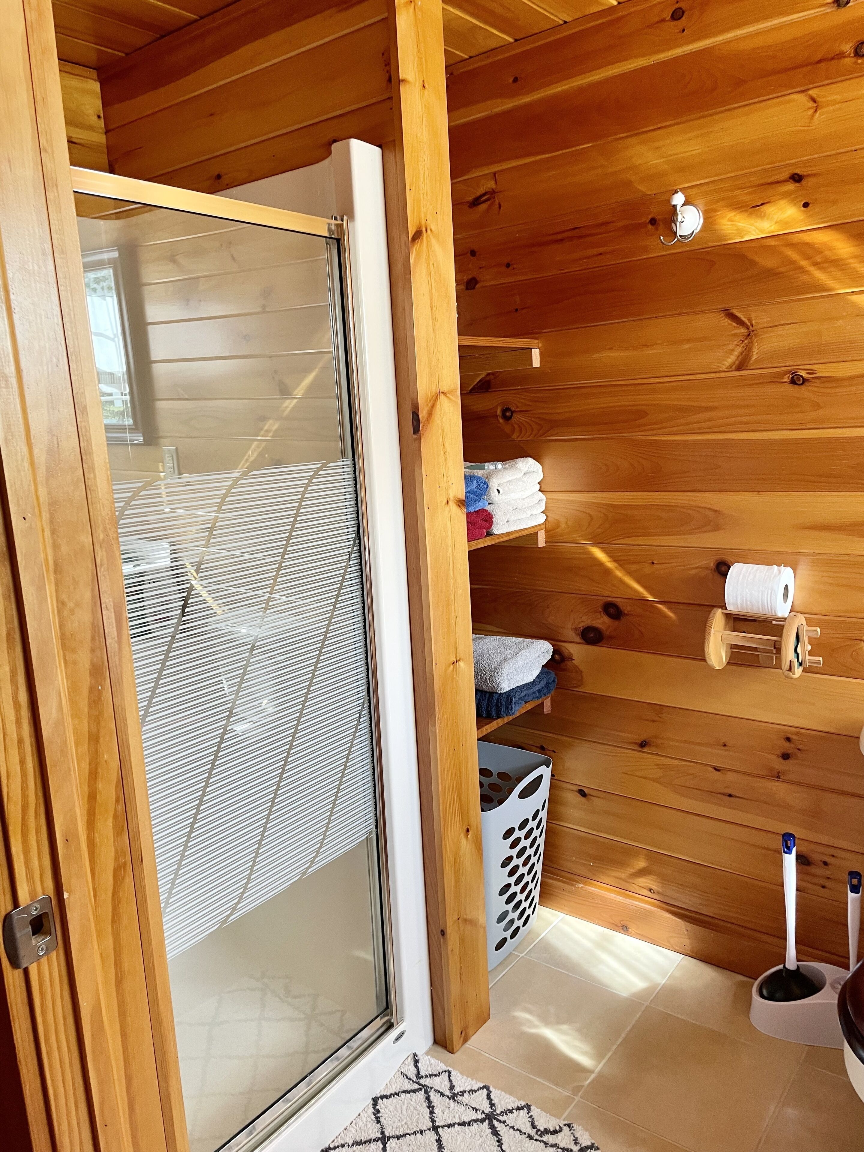 A wooden interior with a sliding glass door, a metal mesh screen, a wooden shelf with towels, a laundry basket, and a white toilet.