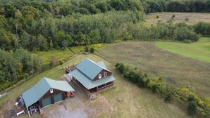 A large barn with a green roof and a smaller adjacent building, surrounded by dense forest and open fields.
