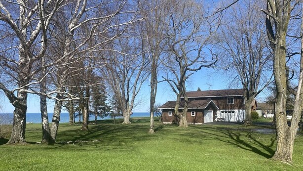 Ein Haus mit Rasen und Bäumen, im Hintergrund ein Gewässer, und ein klarer Himmel.