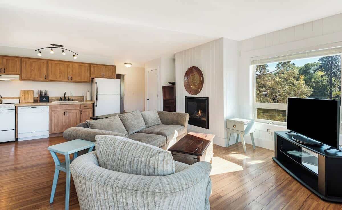 A modern living room with a sofa, coffee table, TV, fireplace, and kitchen area.