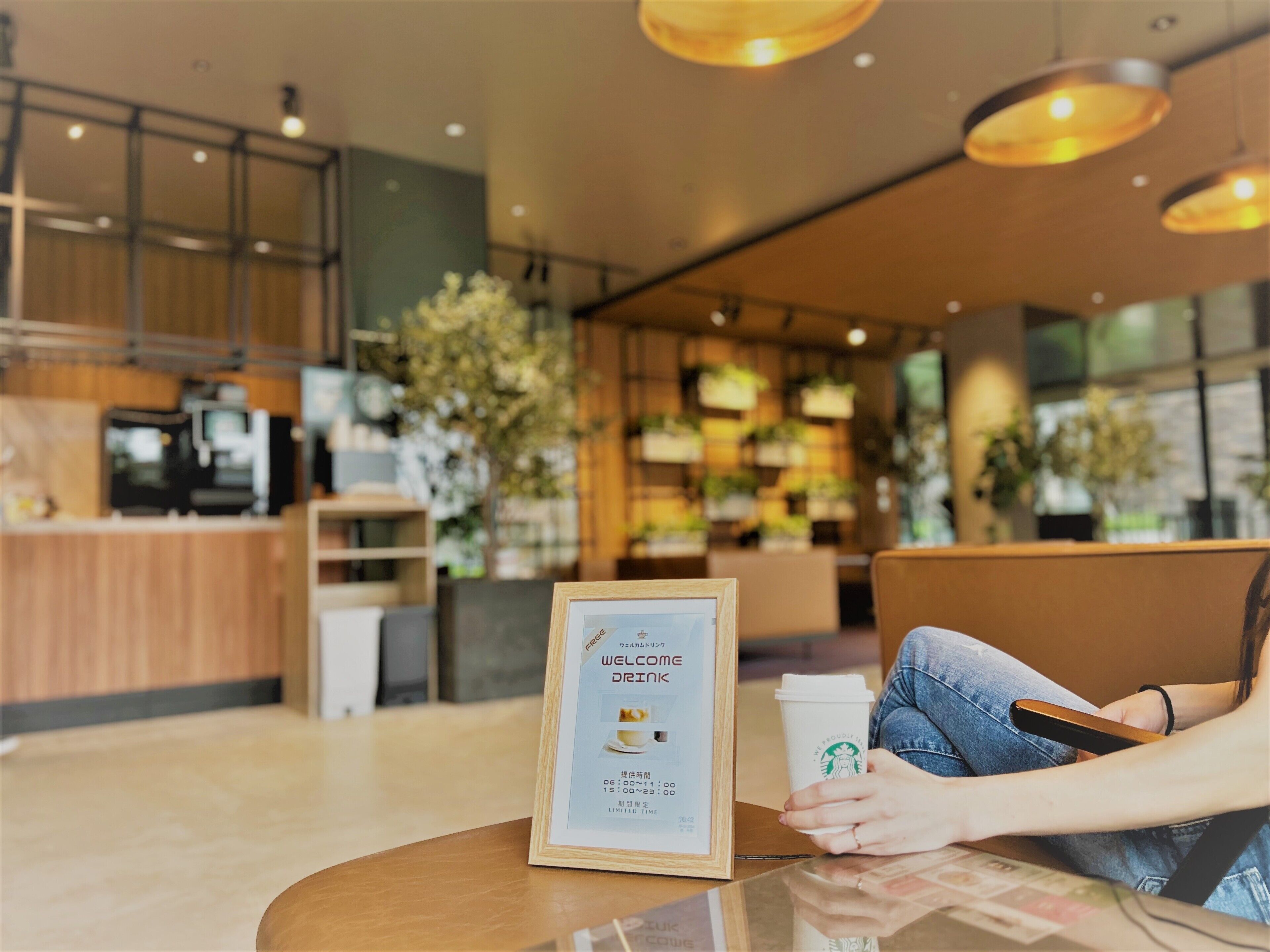 Une personne assise à une table, devant un panneau d’accueil encadré, tenant un gobelet Starbucks et un smartphone.