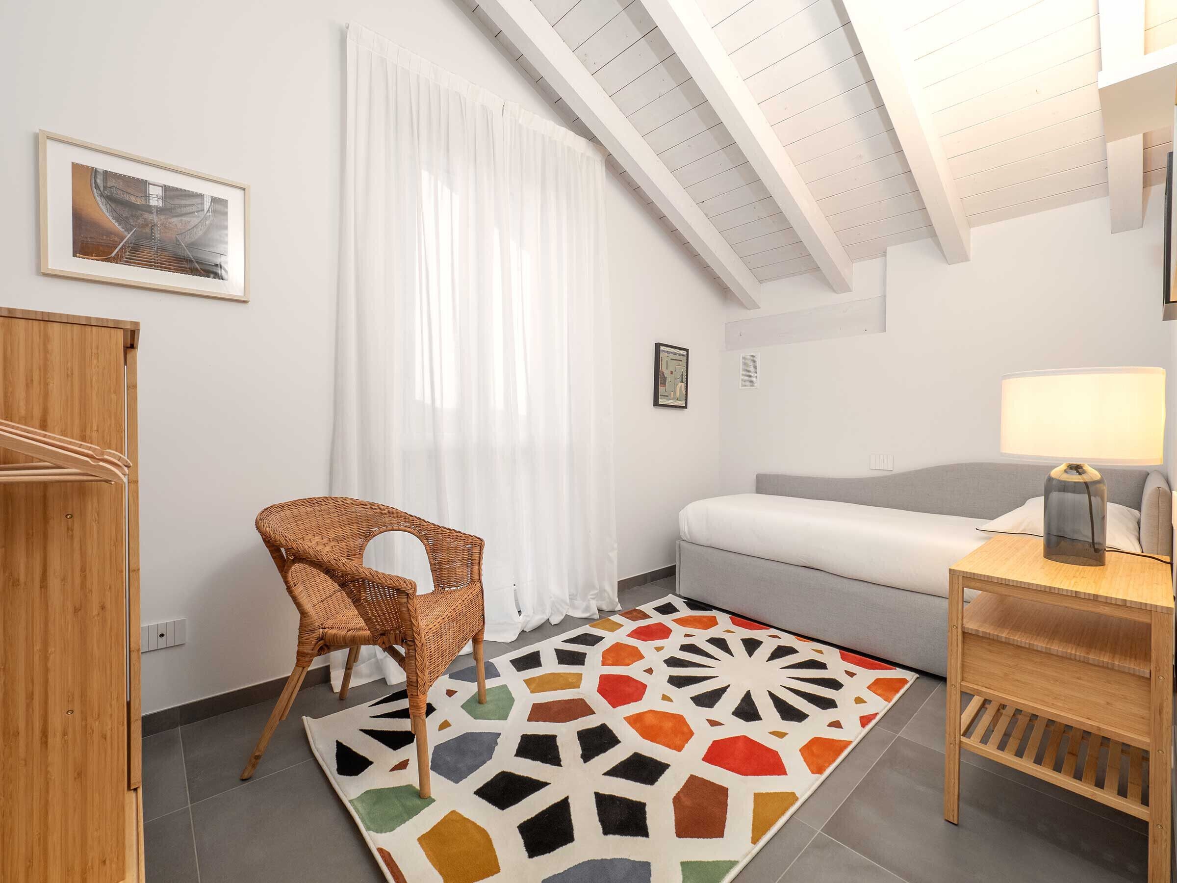 A modern room with a bed, a chair, a table with a lamp, a colorful rug, and a framed picture on the wall.