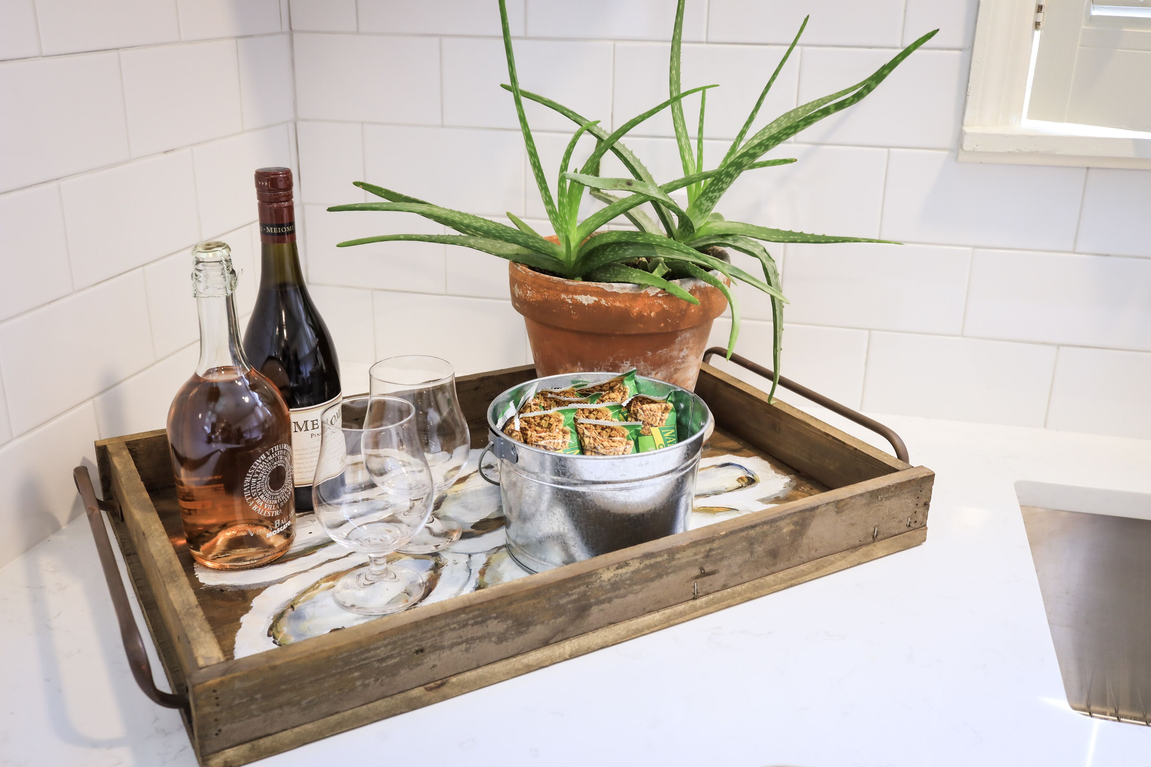 A tray with a bottle, glasses, a plant, and a container of snacks on a kitchen counter.