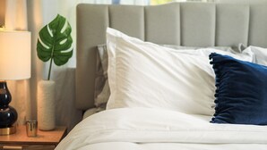 Una cama bien hecha con almohadas blancas y azules, una lámpara de noche y una planta decorativa.