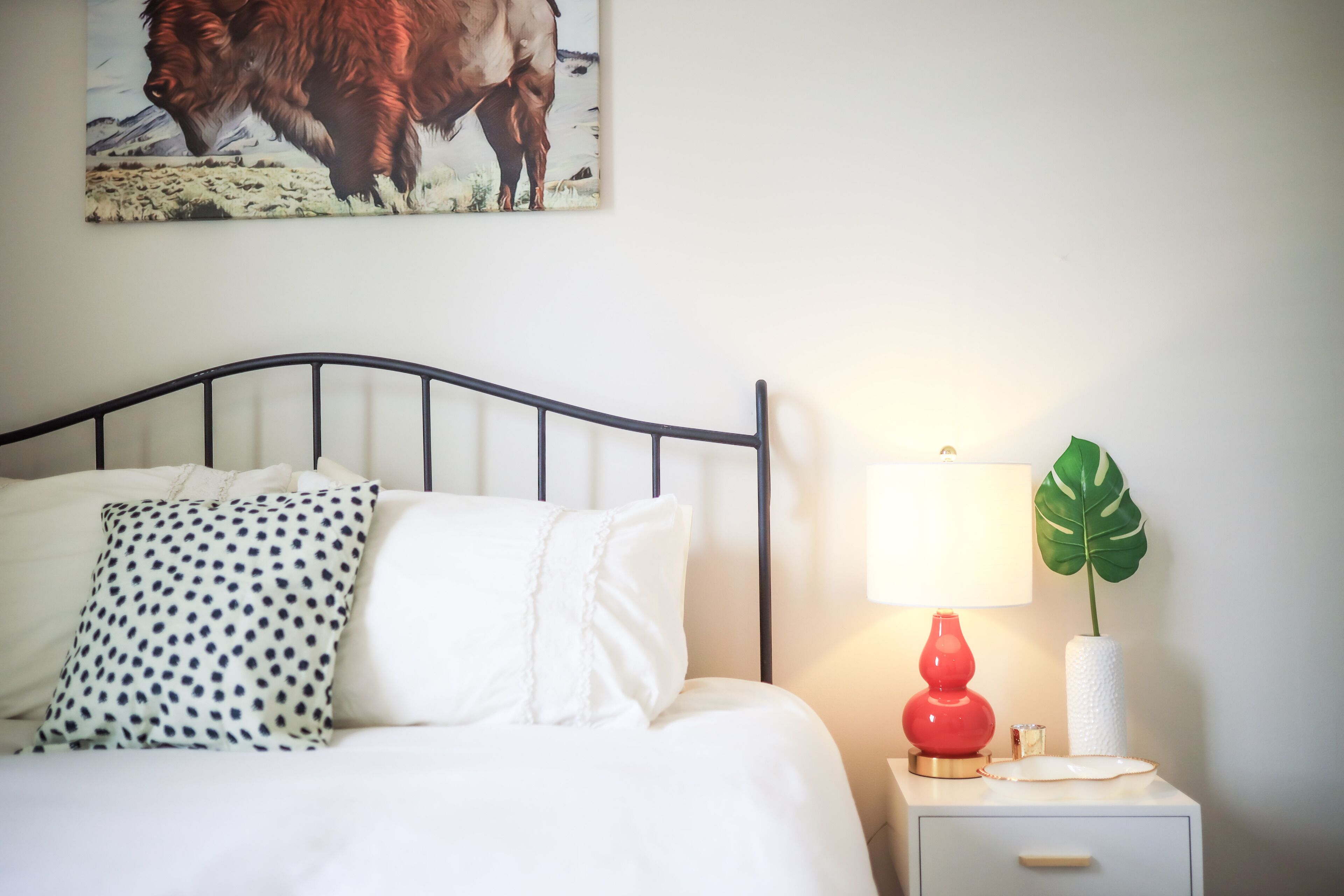 Een netjes opgemaakt bed met witte en stippenbedrukte kussens, een rode lamp en een plant op een nachtkastje.