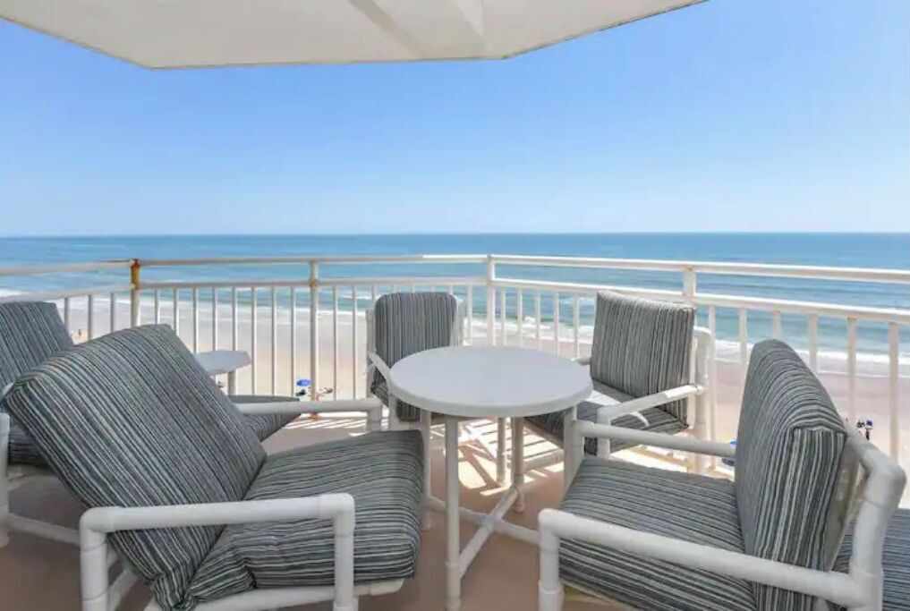 A balcony with striped chairs and a small table, overlooking a beach.