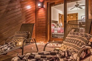 Un cuarto acogedor con techo inclinado de madera, dos sillones reclinables con cojines estampados y un ventanal que da a un dormitorio con cama y ventilador de techo.