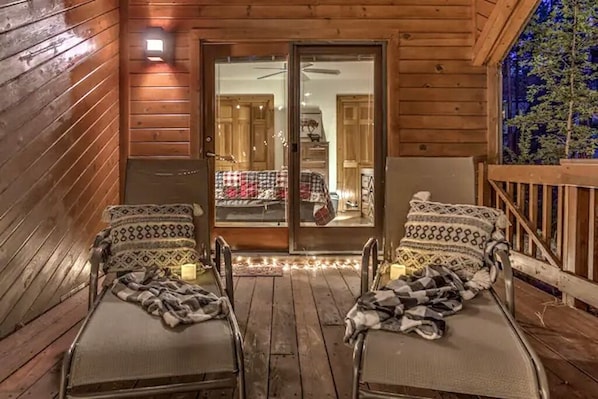 Una acogedora terraza de madera con dos sillas de metal y un banco, una puerta corrediza de vidrio que da a una habitación con una cama y un aparador, y una pequeña lámpara de pared.