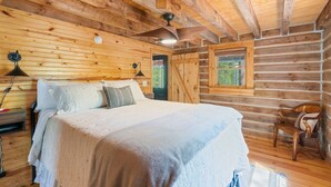 Une chambre dans un chalet en rondins, avec un lit, une chaise, une fenêtre et un ventilateur de plafond.