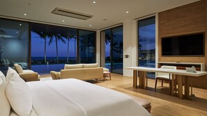 A modern hotel room with a large bed, a desk, and a TV. There is a view of the ocean and palm trees through large windows. - Saba Estate Luxury Villa Bali (Blahbatuh)