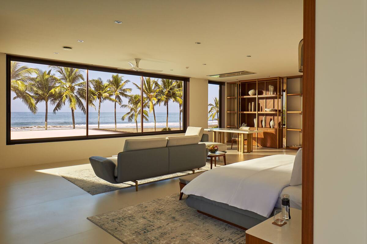 A modern hotel room with a large window overlooking palm trees and the ocean, a bed, a seating area, and a well-equipped bathroom.