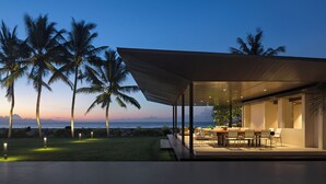A modern outdoor dining area with a glass enclosure, palm trees, and a view of the ocean. - Saba Estate Luxury Villa Bali (Blahbatuh)
