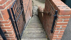 A narrow brick stairway leading to a gated entrance, with a metal gate and a wooden gate on the right.