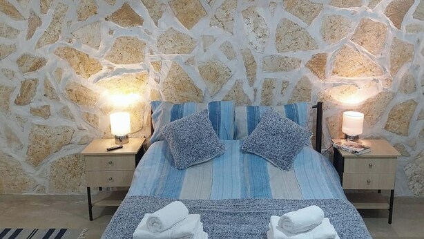 A bed with blue and white bedding, two pillows, and two bedside lamps. The room has a stone wall background. - Lovely Studio in Strinilas with its own pool (Strinilas)
