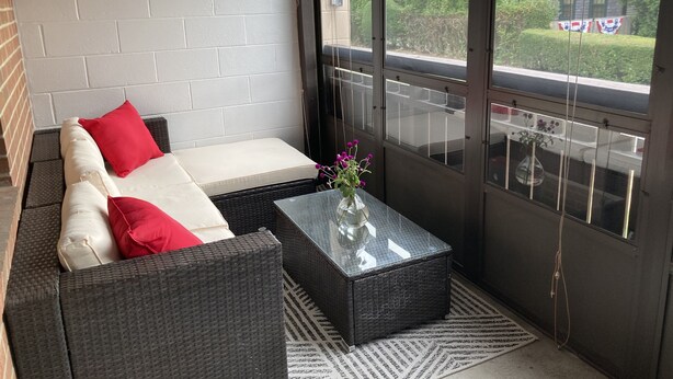 Un balcon avec un canapé en osier, une table basse en verre et une vue sur un immeuble arborant un drapeau.