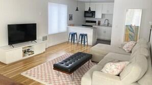 Un salon moderne doté d’un téléviseur à écran plat, d’un canapé, d’un pouf et d’un coin cuisine équipé d’un four à micro-ondes, d’une cuisinière et de tabourets de bar.