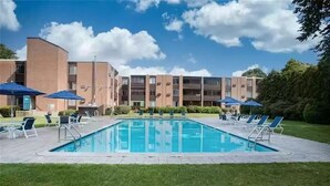 Une piscine entourée de chaises longues et de parasols, le tout bordé par un immeuble à plusieurs étages, sous un ciel dégagé.