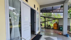 Un espacio al aire libre techado con suelo de baldosas, una silla, una puerta y vista a un jardín con gente y un barril amarillo.