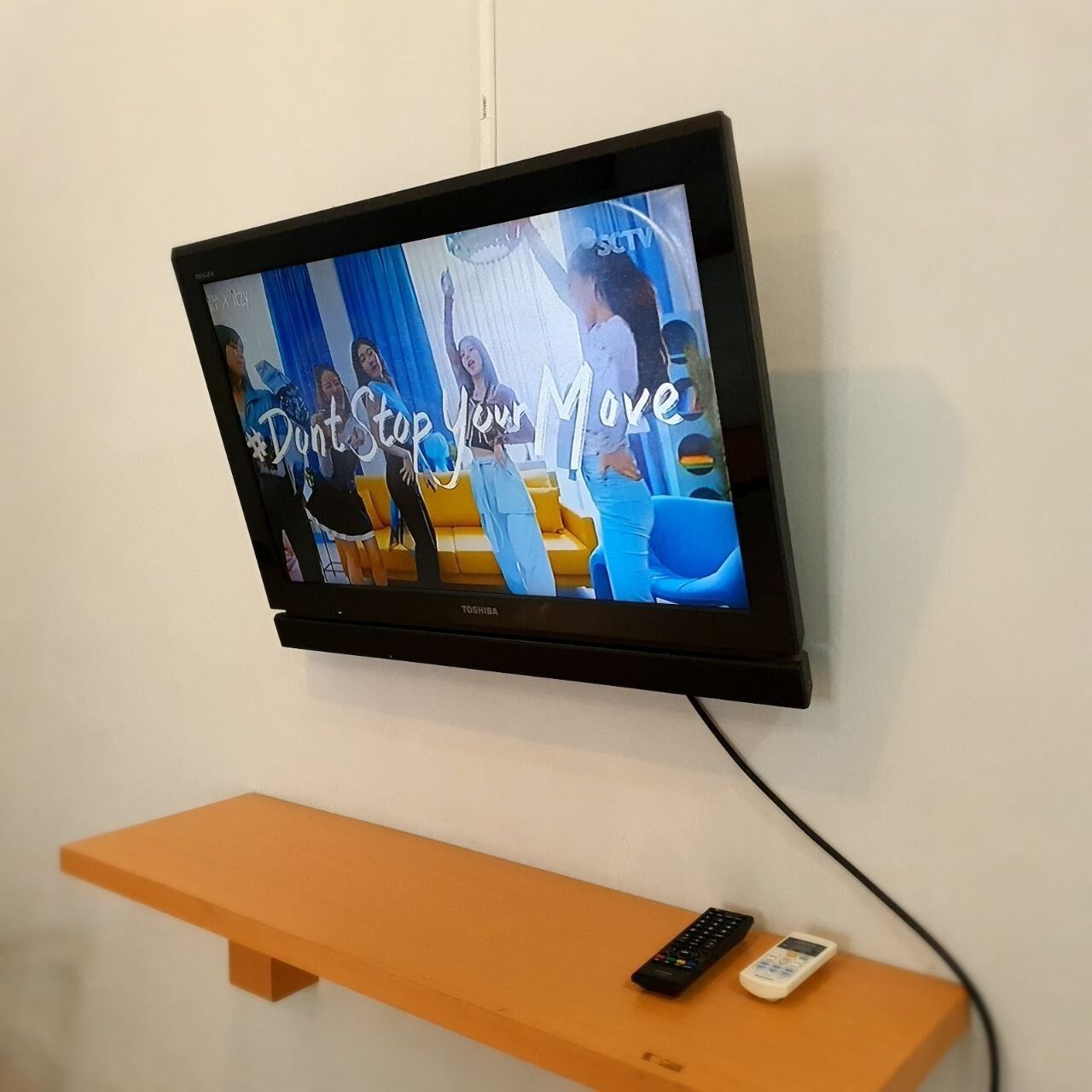 Uma TV de tela plana presa a uma parede branca, exibindo um programa com o texto "Don't Stop Your Move". Abaixo da TV, há uma prateleira de madeira com um controle remoto e um pequeno dispositivo eletrônico.