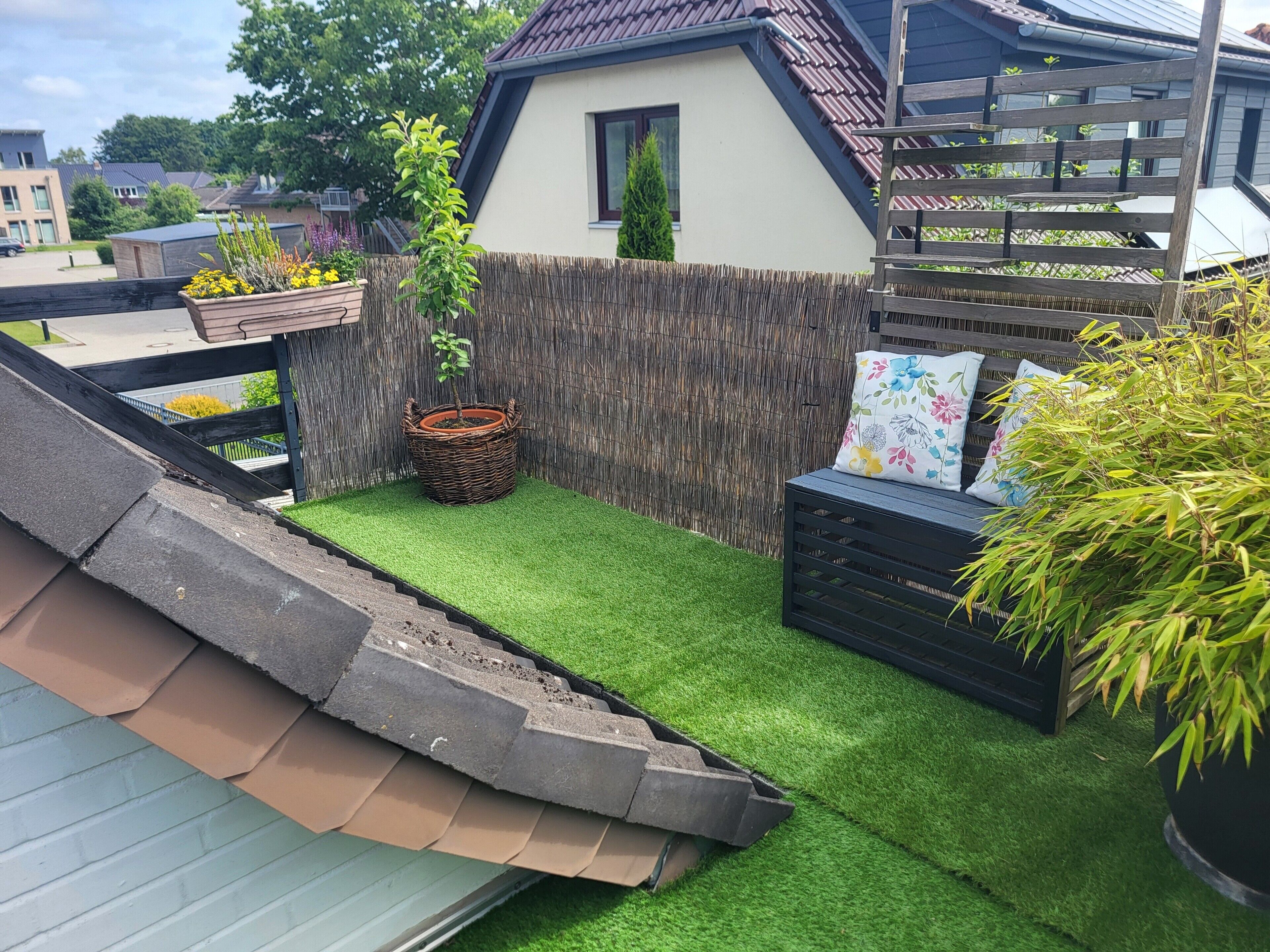 A rooftop garden with artificial grass, a wooden bench, a wicker planter, and a small house-like structure.
