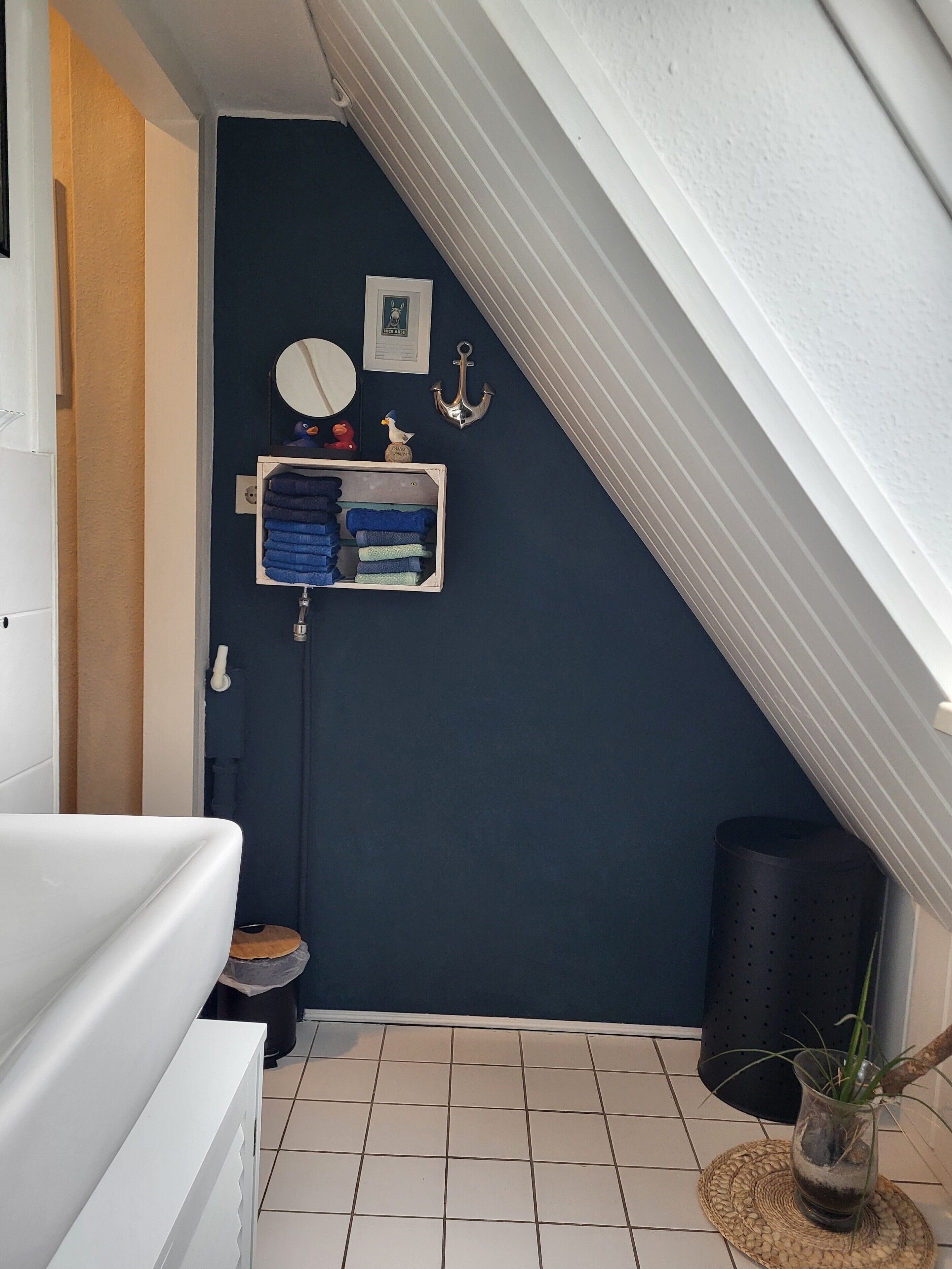 Ein Badezimmer mit dunkelblauer Wand, einer weißen Badewanne, einem kleinen Waschbecken, einer Toilette, einem Handtuchhalter mit gefalteten Handtüchern, einem gerahmten Bild, einer kleinen Anker-Dekoration und einem Topfpflanzen.